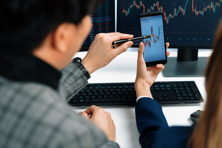 Cropped pointing hand with pen in businessman, checking with holding smartphone by woman investor online website program application with dynamic stock market at home office at back side.の写真素材
