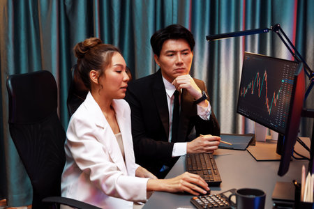 Smiling Asian business traders pointing to dynamic stock exchange data screen profit value online website program at night time at home office. Analyzing currency rate value on data report. Infobahn.の写真素材