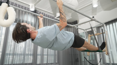Man gripping bar on pilates trapeze table for core strength and upper body workout in a gym. Trainer...