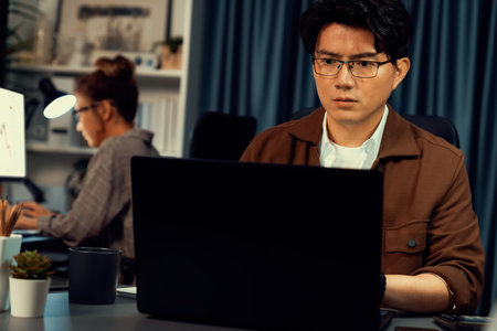 Businessman with stressful face working on laptop waiting email while coworker trading stock exchange investment at late night time at modern office, working on startup company project job. Infobahn.の写真素材