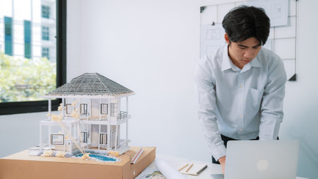 Closeup image of professional architect engineer using laptop analysis data on meeting table with house model and map scatter around at room with blueprint hanged behind. Focus on hand. Immaculate.の写真素材