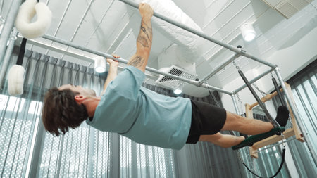 Man gripping bar on pilates trapeze table for core strength and upper body workout in a gym. Trainer hanging inverted back bend on the cadillac for improving flexibility in back and hip. Habituate.の写真素材