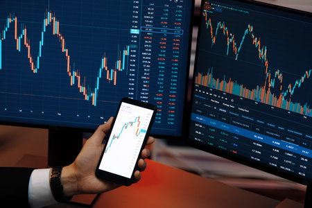 Cropped hand businessman holding smartphone showing dynamic stock market, comparing with financial...