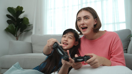 Caucasian mom and daughter sitting and playing game at living room. Happy mother spend time together with smart asian girl while holding joystick ans enjoy playing games. Lifestyle concept. Pedagogy.の写真素材