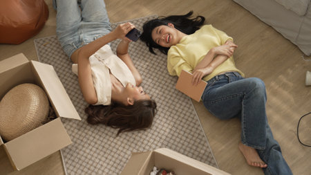 Lover looking at phone while laughing together and lying down at living room floor with cardboard...