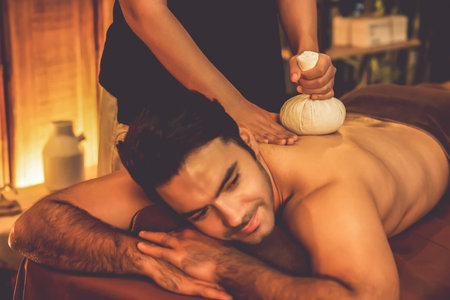 Hot herbal ball spa massage body treatment, masseur gently compresses herb bag on man body. Tranquil and serenity of aromatherapy recreation in warm lighting of candles at spa salon. Quiescentの写真素材