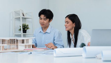 Professional male asian architect using ruler to measure house model length while young beautiful caucasian colleague using laptop to analyzed data on meeting table with house model. Immaculate.の写真素材