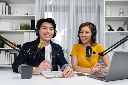 Smiling radio influencer hosts wearing headphones looking camera to pose on social online live streaming with talking topic script note and laptop to listeners at morning at studio record. Infobahn.の写真素材