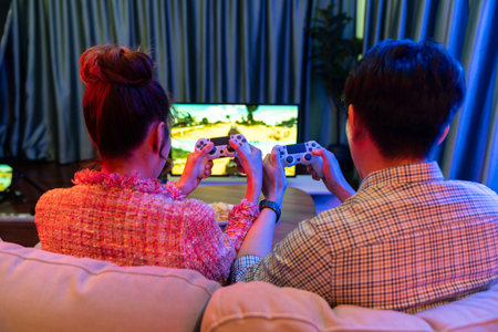 Couple gamer with controlled joystick playing car racing video game together on tv screen with snack at back side view, sitting sofa at neon light colorful living room at modern comfy home. Infobahn.の写真素材