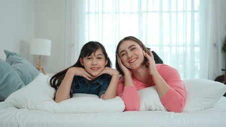 Happy asian daughter and smart mom lie on bed while looking at camera. Energetic family spend time and prepare to sleep together. Cute girl and mom smile to camera show love and affection. Pedagogy.の写真素材