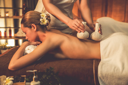 Hot herbal ball spa massage body treatment, masseur gently compresses herb bag on couple customer body. Serenity of aromatherapy recreation in warm lighting of candles at spa salon. Quiescentの写真素材