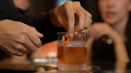 Bartender squeezing orange peel smelling over glass cocktail whiskey putting into Old fashioned on top of rock ice making special drink advertisement alcohol on Paloma and guest background. Vinosity.の写真素材