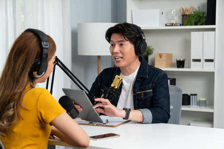 Smiling radio influencer host holding tablet wearing headphones interview woman speaker social media online live streaming channel with talking to share link at morning at studio record. Infobahn.の写真素材