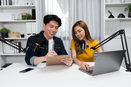 Smiling radio influencer hosts with channel social media online live streaming with talking topic collective question on script note and laptop to listeners at morning time at studio record. Infobahn.の写真素材