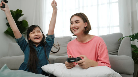 Happy child playing game and winning her mom while sitting at living room. Attractive daughter put...