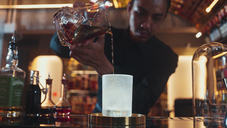 Asian bartender pouring mixed cocktail whiskey into cool glass textured cooling ice drink making special advertisement creative focusing alcohol refresh showcase at luxurious warm light pub. Vinosity.の写真素材