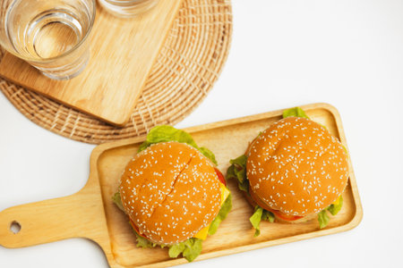 Focusing on double giant hamburger with beef and vegetable meal on square handle wooden board with top angle down view on white background beside drinking glasses water on fast food concept. Infobahn.の写真素材