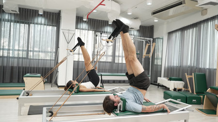 Client performing an inverted leg raise using straps on a reformer machine in a gym. Couple of happy trainer posing reverse crunch for improving core engagement, balance, and flexibility. Habituate.の写真素材