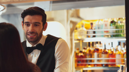 Smiling smart bartender providing excellent service to beautiful guest woman at back side vibrant...