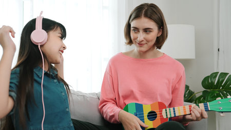 Asian cute girl listening to music from laptop by using headphone while lovely mom playing ukulele while teaching young daughter playing acoustic music. Cute child learning about instrument. Pedagogy.の写真素材