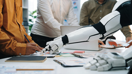 A modern office scene depicting a collaborative workspace where human team members interact with a robotic hand, showcasing the integration of technology and teamwork.Noogenesisの写真素材