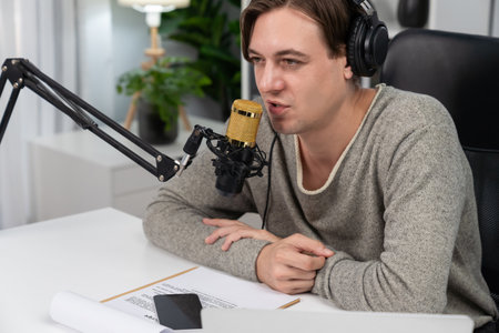 Host channel in smiling smart broadcaster recording, talking show with script and smartphone on desk on live social media, wearing headphones to record video streamer at modern home studio.の写真素材