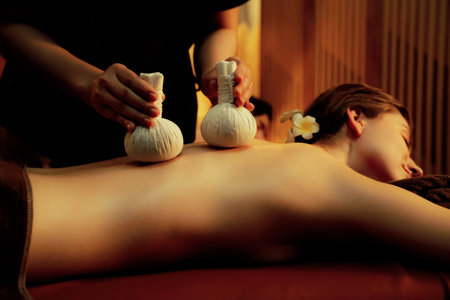 Hot herbal ball spa massage body treatment, masseur gently compresses herb bag on couple customer body. Serenity of aromatherapy recreation in warm lighting of candles at spa salon. Quiescentの写真素材