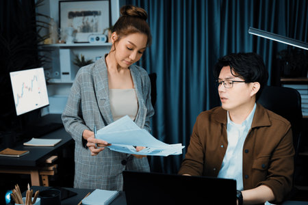 Serious face working on laptop of sales manager with secretary giving overwork paperwork at over late night checking collect sign document for board company keeping database file at office.の写真素材
