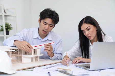 Professional male asian architect using ruler to measure house model length while young beautiful caucasian colleague using laptop to analyzed data on meeting table with house model. Immaculate.の写真素材