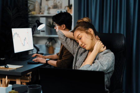 Businesswoman working on desk with stretching arm up and down manner with body health ache of strain overwork while coworker trading stock market on website on pc at late over night time. Infobahn.の写真素材