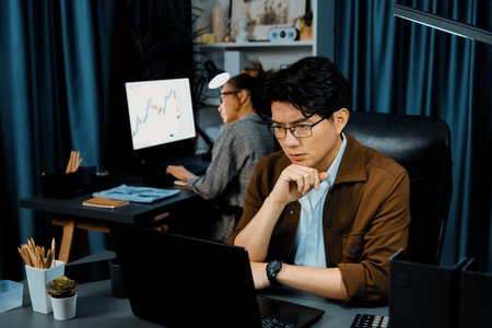 Businessman with stressful face looking on document file deadline while coworker trading stock exchange investment at late night time at modern office working on startup company project job. Infobahn.の写真素材