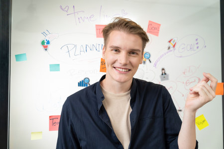 Portrait of happy young businessman wearing blue casual uniform while standing in front of glass board with colorful sticky notes and mind map with confident at creative business meeting. Immaculate.の写真素材