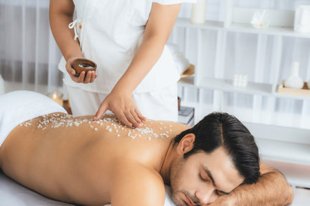 Blissful man customer having exfoliation treatment in luxury spa salon with warmth candle light ambient. Salt scrub beauty treatment in health spa body scrub. Quiescentの写真素材