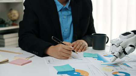A business professional engages in data analysis using a robotic hand, with charts and notes around them, showcasing the blend of humanity and technology in a modern workspace.の写真素材