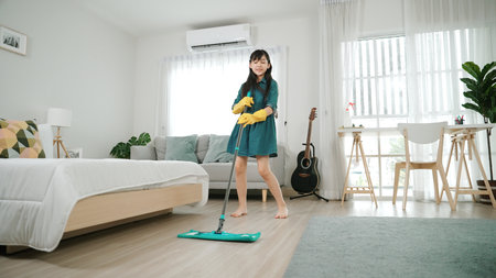 Attractive cute daughter mopping floor while moving to music at modern house. Energetic girl with cleaning gloves doing housework and enjoy cleaning bedroom while singing a songs. Lifestyle. Pedagogy.の写真素材