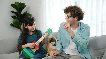 Back view of happy father looking at smart daughter playing ukulele warmly and singing song. Happy dad motivated and encouraged american school girl enjoy playing guitar. Family recreation. Pedagogy.の写真素材