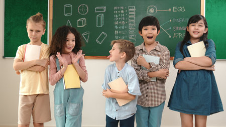 Multicultural elementary student in casual cloth looking at camera while standing at classroom. Group of cute diverse children hold notebook and smiling to camera at blackboard in classroom. Pedagogy.の写真素材