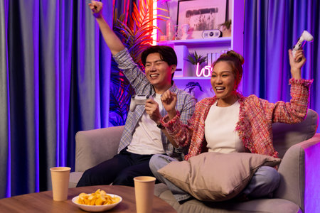 Couple gamer raising fist up holding joysticks be winner together complete level sitting sofa...