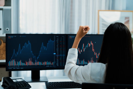 Successful young Asian businesswoman earning highest profit with fist up in exchange stock market on dynamic business graph on pc screens at modern office, analyzing financial technology. Stratagem.の写真素材