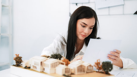 Closeup of young beautiful architect compares blueprints with architectural model with house model and architectural equipment placed on table at meeting room. Creative design concept. Immaculate.の写真素材