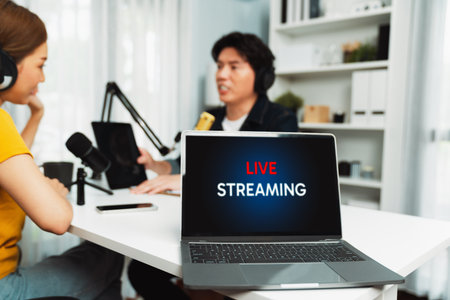 Selective focusing on live streaming laptop screen on blurry background of smiling radio influencer hosts wearing headphone with channel social media online holding tablet at studio record. Infobahn.の写真素材