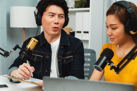 Smiling radio influencer hosts wearing headphones with channel social media online live streaming with talking topic on script note and laptop to listeners at morning time at studio record. Infobahn.の写真素材