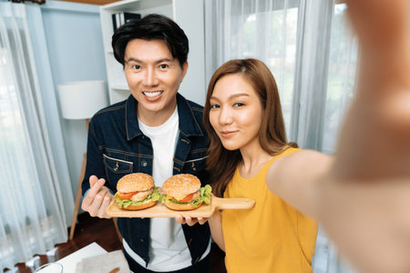 Smiling host chef influencers taking photo with smartphone by selfie straight view holding double hamburger on cooking show special easy dish homemade on social media channel live streaming.の写真素材