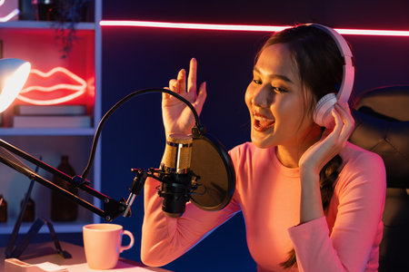 Host channel of young beautiful Asian broadcaster listening the music on social media with listeners, wearing pastel headphones, using microphone for singing at neon light indoor studio. Stratagem.の写真素材