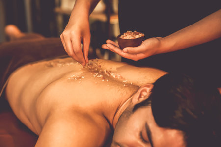 Man customer having exfoliation treatment in luxury spa salon with warmth candle light ambient. Salt scrub beauty treatment in Health spa body scrub. Quiescentの写真素材