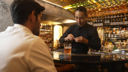 Bartender squeezing orange peel smelling over glass cocktail whiskey putting into Old fashioned on top of rock ice making special presenting to VIP customer at luxurious counter bar at club.の写真素材