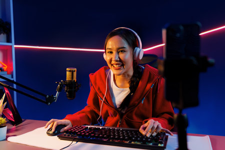 Host channel of smiling beautiful Asian girl streamer playing online game wearing headphone talking with viewers media online recording phone. Esport skilled team players in neon blue room. Stratagem.の写真素材