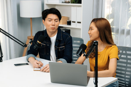 Smiling radio influencer hosts with channel social media online live streaming with talking topic collective question on script note and laptop to listeners at morning time at studio record. Infobahn.の写真素材