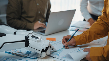 A robotic arm collaborates with human colleagues at a modern office workstation, analyzing business documents and exploring innovative strategies for productivity.の写真素材