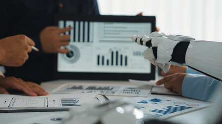 A modern business meeting scene showcasing teamwork with human hands and a robotic arm analyzing dynamic graphs and charts on a digital tablet, emphasizing innovation.Noogenesisの写真素材